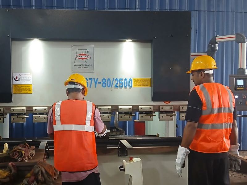 The press brake bay. The 80 tonne by 2500 mm CNC press brake. Both operators in helmet, high-visibility vest and gloves.