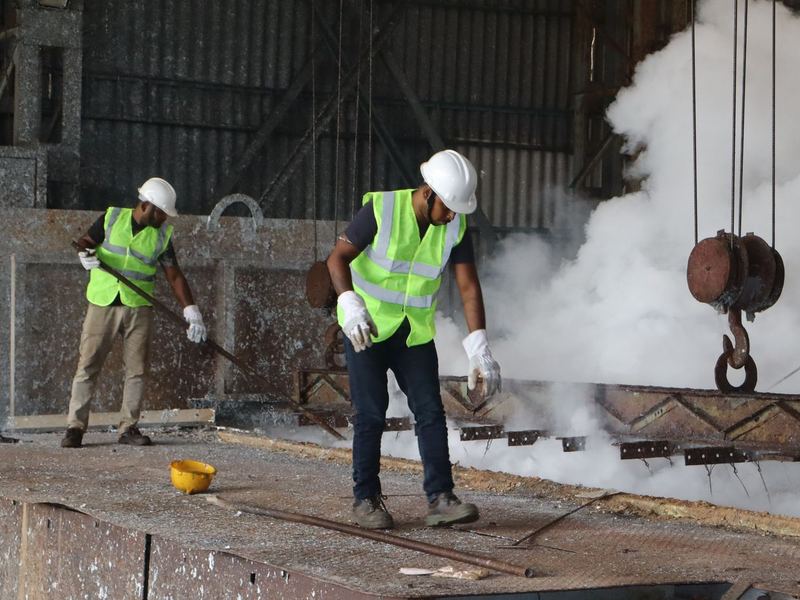 Operators at the zinc kettle. Working the kettle at 450 degC. Helmets, high-visibility vests and gloves on the kettle floor; the lifting beam and jig are our own.